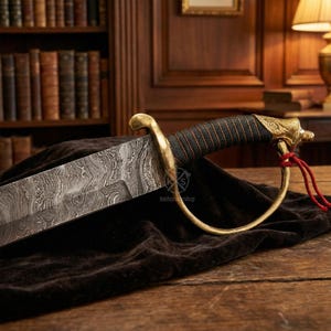 May include: A decorative saber with a patterned blade and a gold-colored handle. The handle is wrapped in black material and features a detailed design. A red cord is attached to the handle. The saber rests on a dark cloth.