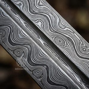 May include: Close-up of a metal blade with a distinctive pattern. The blade's surface features a swirling, layered design in shades of gray and silver, creating a textured appearance. The design resembles wood grain or water ripples.