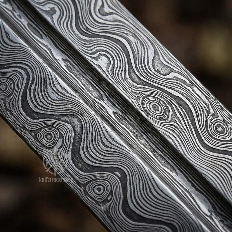 May include: Close-up of a metal blade with a distinctive pattern. The blade's surface features a swirling, layered design in shades of gray and silver, creating a textured appearance. The design resembles wood grain or water ripples.