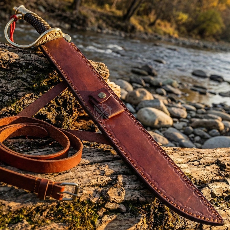 May include: A brown leather sword scabbard with a matching belt and a decorative sword. The sword has a gold-colored hilt and a dark brown handle. The scabbard is resting on a log near a river.