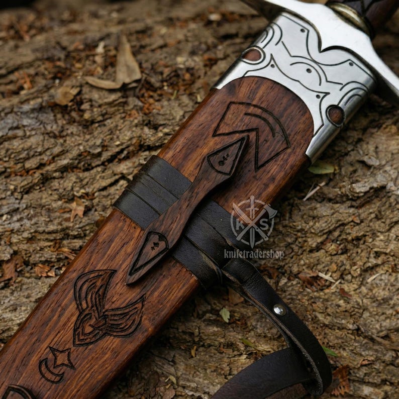 May include: Close-up of a sword in a wooden sheath. The sheath is dark brown with carved designs and a leather strap. The sword's hilt is visible, with a silver guard and a dark brown handle. The text "knifetradershop" is visible.