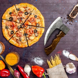 May include: A pizza with cheese, pepperoni, olives, and tomato slices is cut into eight slices. The pizza is on a dark purple surface with a large, curved knife, a bag of spaghetti, and other ingredients.