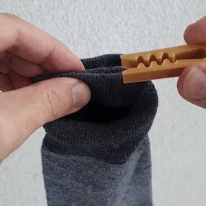 以下が含まれることがあります： 2つの手で持たれた灰色の靴下のクローズアップ。木製の靴下クリップが付いています。クリップは薄茶色で波状のデザインです。靴下は濃い灰色です。