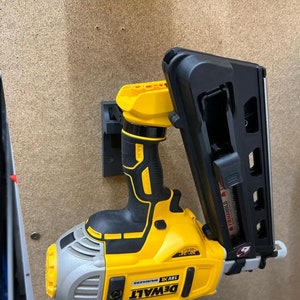 Op de afbeelding: Een gele en zwarte DeWalt 18V X Brushless 30-graden afwerknagelpistool is gemonteerd op een zwarte wandbevestiging. Het spijkerpistool heeft een zwart en zilveren magazijn en een zwarte trekker.