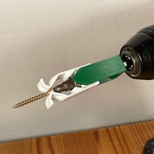 Puede incluir: Un ancla de pared de plástico blanco con un tornillo unido a un taladro eléctrico negro y verde.