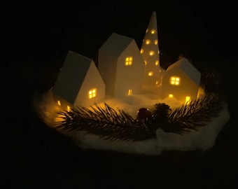 May include: A white ceramic village scene with a tall, pointed tree in the center. The houses and tree have small, warm lights inside, creating a cozy and festive atmosphere. The scene is surrounded by a bed of white faux snow and green pine branches.