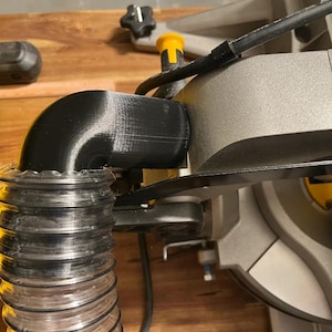 May include: A black plastic dust collection adapter with a flexible hose attached to it. The adapter is connected to a grey and black power tool.