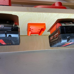 Op de afbeelding: Een close-up van een houten plank met drie zwarte en rode accu's voor elektrisch gereedschap die hangen aan oranje kunststof houders. De accu's zijn gelabeld met "M18 CP3.0", "M18 REDLITHIUM XC5.0" en "M18 FUEL 3.0".