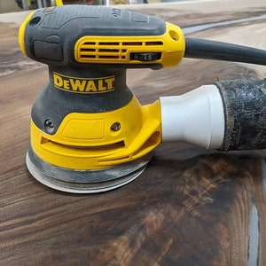 Puede incluir: Una lijadora orbital aleatoria DeWalt amarilla y negra con una manguera de recolección de polvo. La lijadora está sobre una superficie de madera marrón oscura.
