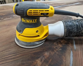 May include: A yellow and black DeWalt random orbit sander with a dust collection hose attached. The sander is sitting on a dark brown wooden surface.