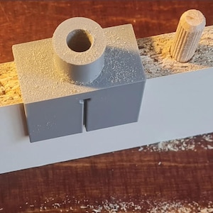 Puede incluir: Una plantilla de clavija de plástico gris colocada sobre una tabla blanca y de madera. Una clavija de madera está al lado de la plantilla. El aserrín está esparcido alrededor de la plantilla y la tabla. La plantilla tiene una parte superior cilíndrica y una base rectangular con una ranura.