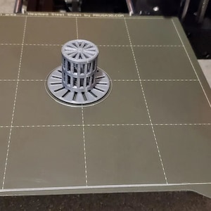 May include: A gray 3D printed object with a circular base and a cylindrical top with a grid pattern. The object is sitting on a gray textured surface with a grid pattern. The text "ORIGINAL PRUSA MINI" is visible on the surface.