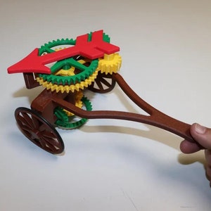Op de afbeelding: Een bruine houten speelgoedkar met een handvat en een set kleurrijke tandwielen. De tandwielen zijn rood, groen en geel. De kar heeft één wiel aan de voorkant en een groter wiel aan de achterkant. De tandwielen zijn verbonden met een rode pijl die draait wanneer het handvat wordt gedraaid.