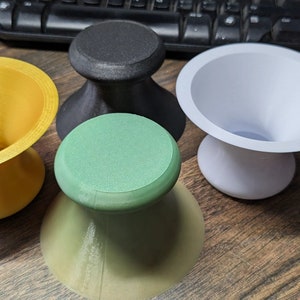 Könnte beinhalten: Vier unterschiedlich farbige Keramik- oder Plastik-Schalen mit einer breiten Oberseite und einem schmalen Boden. Die Schalen sind schwarz, weiß, gold und grün.