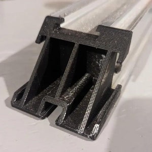 Könnte beinhalten: Nahaufnahme einer schwarzen Kunststoff-Eckhalterung mit strukturierter Oberfläche. Die Halterung hat drei Schlitze und ist so konzipiert, dass sie zwei Materialstücke miteinander verbindet.
