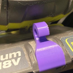 Könnte beinhalten: Ein lilafarbener Plastikhaken, der an einem schwarz-gelben Elektrowerkzeug befestigt ist. Das Werkzeug trägt den Schriftzug "18V".
