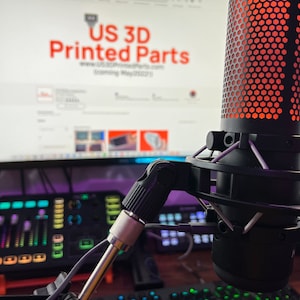 Puede incluir: Un micrófono negro con una rejilla de malla roja y un filtro antipop. El micrófono está montado en un brazo de extensión y está colocado frente a un monitor de computadora. El monitor muestra un sitio web para "US 3D Printed Parts".