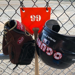 May include: A brown leather baseball glove with a red logo, a black baseball helmet with the word "WIN" in white letters and a red baseball, and a wooden baseball bat with the word "Slugger" printed on it. The items are hanging on a chain link fence with a bright orange sign that says "99" hanging above them.