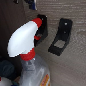 Puede incluir: Soporte negro para botella con pulverizador montado en una superficie de madera. El soporte es de metal y tiene un diseño curvado para sujetar de forma segura una botella con pulverizador. La botella tiene una boquilla blanca y roja.