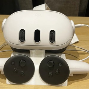 Puede incluir: Un auricular de realidad virtual blanco con dos mandos blancos. El auricular tiene tres sensores negros en la parte delantera. Los mandos tienen botones negros y una palanca de mando negra.