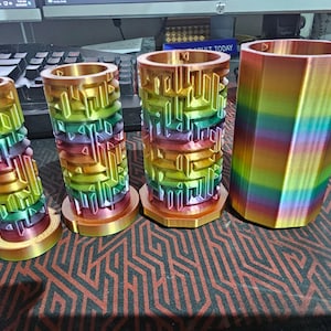 May include: Four colourful 3D printed cylindrical containers with a rainbow pattern. The containers are different sizes and are made of a smooth, glossy plastic.