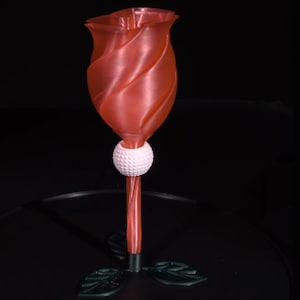 Puede incluir: Un vaso con forma de rosa impreso en 3D con una bola de golf blanca en la base. El tallo es rojo y las hojas son verdes.