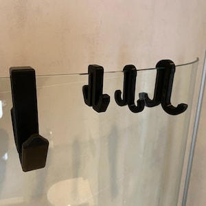 Puede incluir: Cuatro ganchos de plástico negro montados en una puerta de ducha de vidrio. Los ganchos están diseñados para sujetar artículos como toallas o batas.