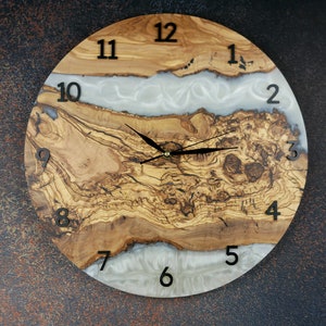 Peut inclure: Une horloge ronde en bois avec un incrustation de résine blanche. Le cadran de l'horloge a des chiffres romains noirs et des aiguilles noires. Le bois a une couleur naturelle brun et gris avec un motif de grain unique.