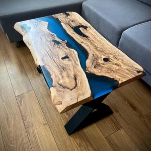 Op de afbeelding: Een rechthoekige salontafel met een houten blad met natuurlijke boomrand, een opvallende houtnerf en een centraal rivierachtig inlegwerk van doorschijnende blauwe epoxyhars, ondersteund door een zwart metalen onderstel in een Z-vorm.