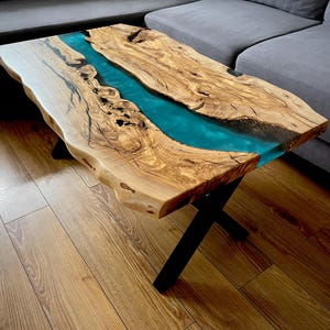 Può includere: Un tavolino da caffè in legno con un piano a bordo vivo e un fiume di resina epossidica blu turchese che attraversa il centro. Il tavolo ha una base a X in metallo nero.