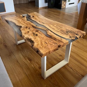 Op de afbeelding: Een rechthoekige salontafel met een olijfhouten blad met natuurlijke boomrand, een opvallende houtnerf en een centrale inleg van doorschijnende grijze hars. Het tafelblad rust op twee stevige, U-vormige witte metalen poten.