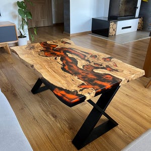 Könnte beinhalten: Ein Couchtisch mit natürlicher Kante und einzigartigem Design. Die Tischplatte aus hellem Holz hat einen Fluss aus orangefarbenem und schwarzem Kunstharz. Schwarze Metallbeine in X-Form verleihen einen modernen Look. Maße in Zentimetern.