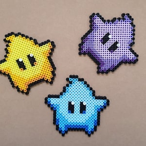 May include: Three pixel art stars in yellow, purple, and blue. Each star has a black outline and a smiling face. The stars are made of small, colorful beads, and are likely handmade. The background is a neutral tan color.