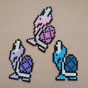 Könnte beinhalten: Drei bunte Perlenfiguren, jede mit einem anderen Design. Die erste Figur ist eine lila und weiße Schnecke mit einer lila Muschel. Die zweite Figur ist eine blaue und weiße Schnecke mit einer blauen Muschel. Die dritte Figur ist ein rosa und weißer Schmetterling.