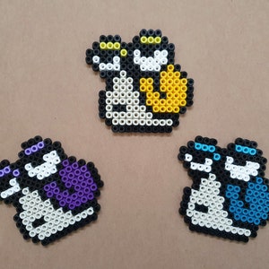 Könnte beinhalten: Drei Pixel-Art-Charaktere aus bunten Perlen. Die Figuren sind weiß mit schwarzen Umrissen und haben verschiedene farbige Akzente. Ein Charakter hat gelbe Akzente, einer hat lila Akzente und einer hat blaue Akzente.