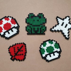 Könnte beinhalten: Fünf verschiedene Pixel-Art-Designs aus bunten Perlen. Die Designs umfassen einen roten und weißen Pilz, einen grünen Pilz, einen grünen Frosch, einen weißen Stern und eine rote Blume.