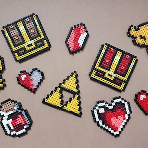 Könnte beinhalten: Eine Sammlung von Pixel-Art-Bastelarbeiten mit Charakteren und Gegenständen aus der Videospielserie The Legend of Zelda. Die Bastelarbeiten sind aus bunten Perlen hergestellt und beinhalten eine Truhe, einen Schlüssel, einen Herzbehälter, einen Trank und das Triforce-Symbol.