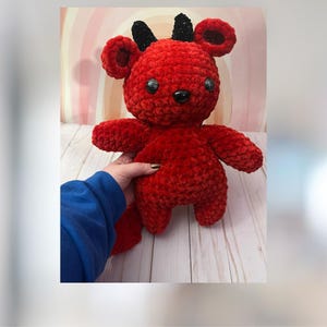 May include: Red crocheted plush toy with black horns and black eyes. The toy is standing on a wooden surface.