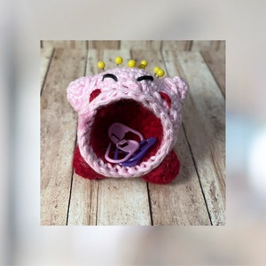 Puede incluir: Un amigurumi de crochet rosa y rojo con botones amarillos para los ojos y una corona de cuentas amarillas. El amigurumi tiene una gran boca abierta y está lleno de marcadores de punto de plástico de colores.