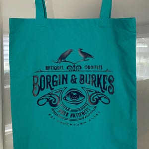 Könnte beinhalten: Eine türkisfarbene Tragetasche mit einem schwarz-weißen Logo, auf dem "Borgin & Burkes" steht. Über dem Logo steht der Text "Antiquitäten & Kuriositäten" und unter dem Logo "Dark Artifacts 37 Knockturn Alley". Zwei schwarze Raben sitzen auf einem Ast über dem Logo.