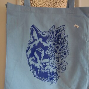 Könnte beinhalten: Eine hellblaue Tragetasche mit einem blauen Wolfskopf-Design. Der Wolfskopf ist von blauen Blumen umgeben. Die Tasche hat zwei blaue Henkel.