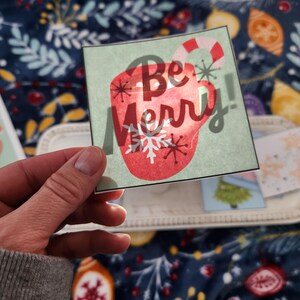 Puede incluir: Una tarjeta verde y blanca con una taza roja y el texto "Be Merry!" impreso en ella. La taza tiene un bastón de caramelo y copos de nieve.