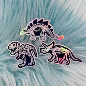 May include: Three iridescent dinosaur skeleton stickers on a blue fluffy background. The stickers are of a Tyrannosaurus Rex, a Triceratops, and a Stegosaurus.