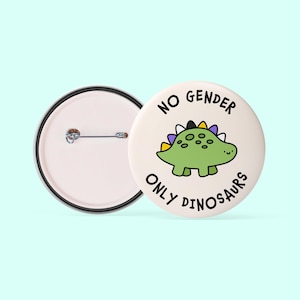 May include: A white button featuring a green dinosaur with purple, yellow and black stripes on its back. The text "NO GENDER ONLY DINOSAURS" is written around the dinosaur.