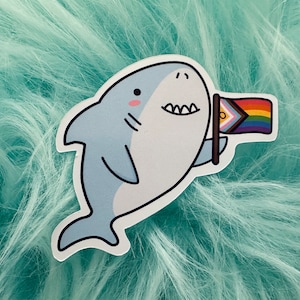May include: A cartoon shark sticker with a friendly expression, holding a rainbow pride flag with a yellow triangle in the top left corner. The sticker is on a light blue fuzzy background.