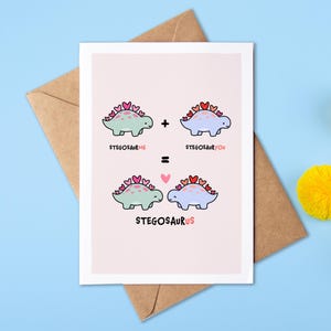 May include: A pink card with a cute illustration of two blue and green dinosaurs with hearts on their backs. The text on the card reads "Stegosaurus Me + Stegosaurus You = Stegosaurus Us".