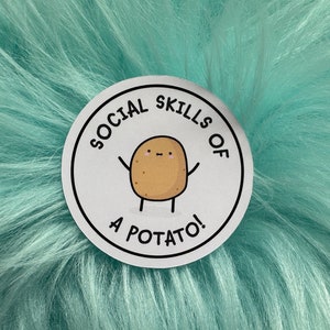 May include: A circular white sticker featuring a cartoon illustration of a tan potato with a simple face and stick arms and legs. The text "SOCIAL SKILLS OF A POTATO!" is printed in black capital letters in a circular arc around the potato.