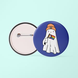 May include: A blue button featuring a white ghost wearing an orange hat and holding a rainbow flag. The reverse of the button is plain.
