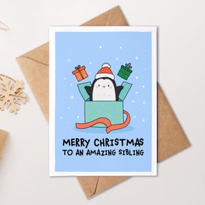 May include: A Christmas card featuring a cartoon penguin in a Santa hat emerging from a gift box. The card reads "MERRY CHRISTMAS TO AN AMAZING SIBLING" on a light blue background with snowflakes. It rests on a brown envelope.