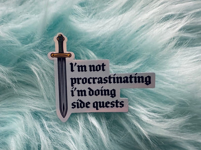 Puede incluir: Una pegatina con una espada y el texto "I'm not procrastinating I'm doing side quests". La pegatina tiene un borde plateado y est&aacute; sobre un fondo azul claro y esponjoso.